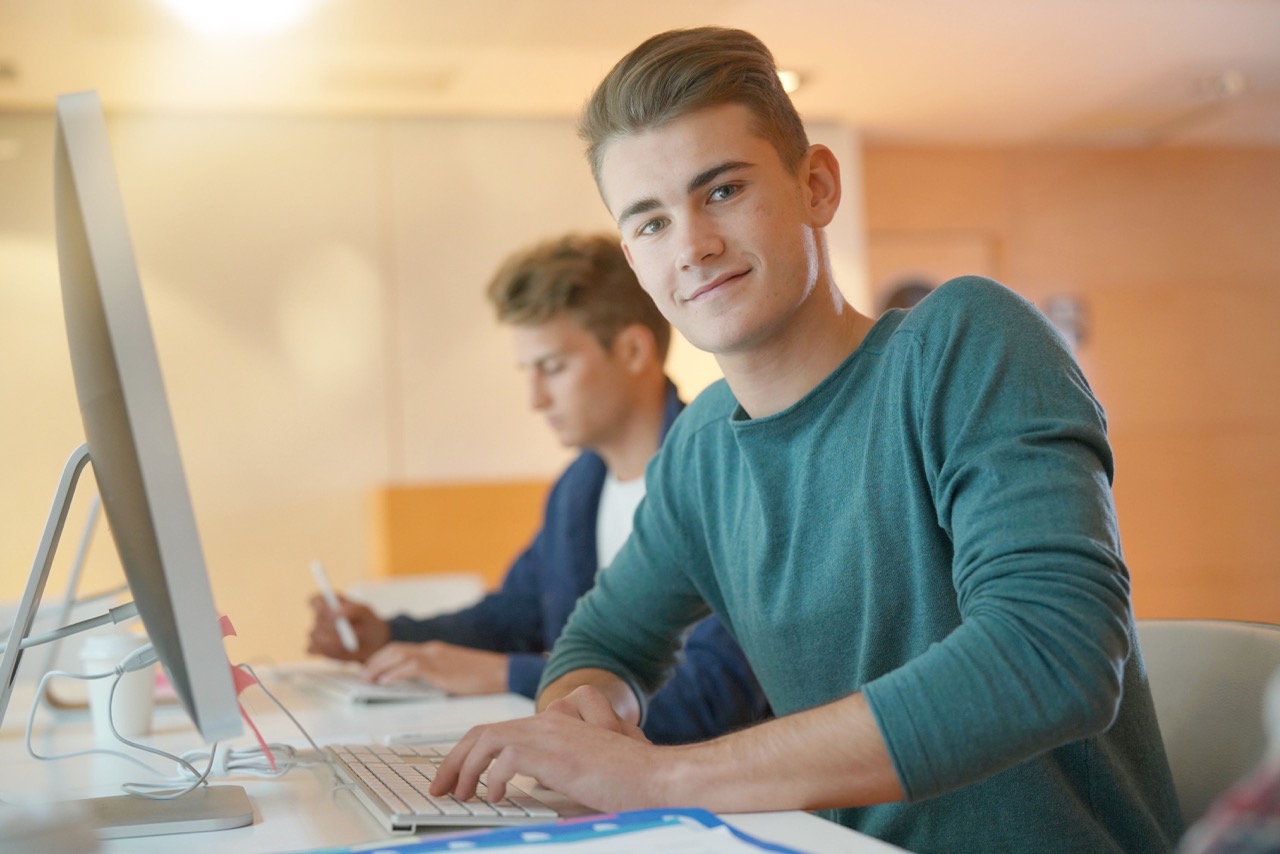 Young man sitting at desk smiling, wearing green long sleeve shirt