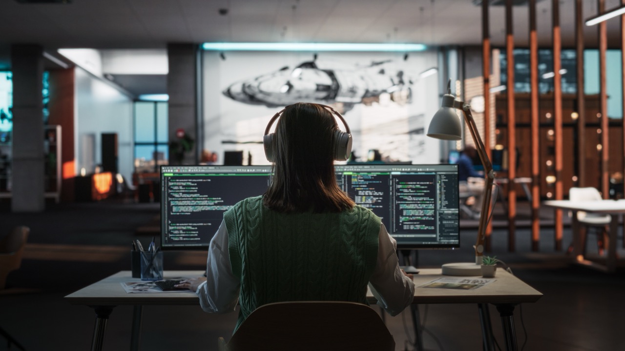 Woman sitting at computer with headphones and screens open with code displayed